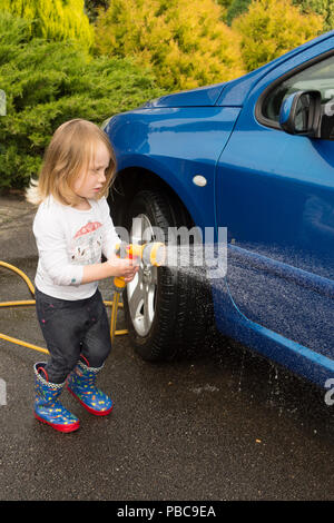Drei Jahre altes Mädchen mit Schlauch helfen blaues Auto zu waschen, Spritzen, Karosserie, Großbritannien. Stockfoto