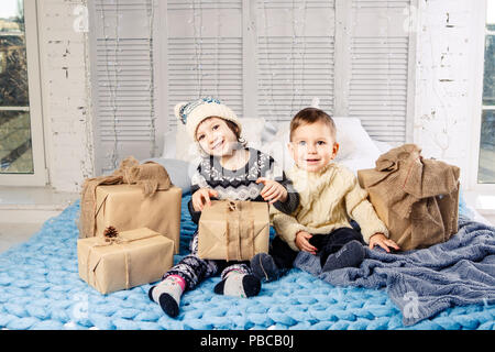 Thema Weihnachten Morgen. Zwei untergeordnete Kaukasischen Junge und Mädchen Bruder und Schwester sind auf dem Bett zu sitzen in einer Umarmung mit einem Lächeln und Freude sind die Eröffnung neuer Stockfoto