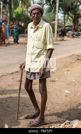 Belathur, Karnataka, Indien - November 1, 2013: Schlank alter Mann mit gelben Hemd und braune Hosen geht mit Stock auf Feldweg. Stockfoto