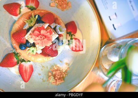 Ansicht von oben heiße Pfanne Kuchen gekrönt mit frischen Erdbeeren, Blaubeeren, Wabe und frische Blumen für die Dekoration auf weißem Teller Stockfoto