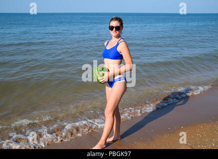 Die Blondine in Sonnenbrille steht an der Küste und hält eine ganze Wassermelone in den Händen. Eine reife Wassermelone in den Händen einer Frau Stockfoto