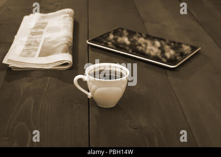 Auf dem braunen Holztisch sind Cap mit Untertasse, Zeitung und Tablet-Konzept Kaffeepause in Sepia Farbe. Stockfoto