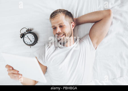 Stattlicher Mann mit einem digitalen Tablette im Bett zu Hause Stockfoto