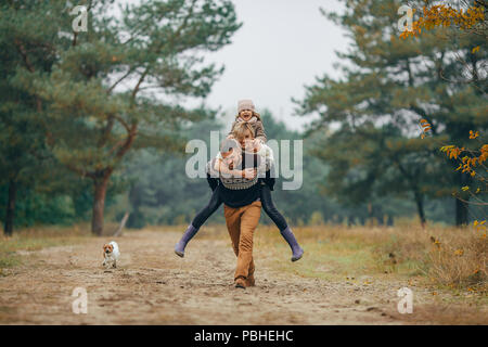 Glückliche Familie hat Spaß und Mann seine Frau und seine Tochter trägt auf seinem Rücken im Wald Weg neben Hund beim Spaziergang im Wald. Stockfoto