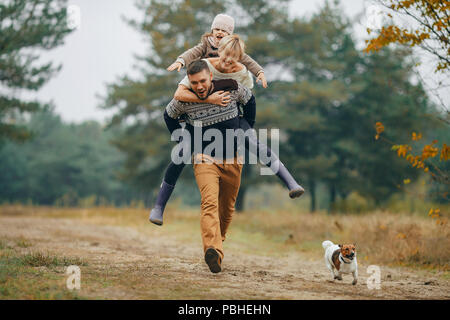 Glückliche Familie hat Spaß und Mann seine Frau und seine Tochter trägt auf seinem Rücken im Wald Weg neben Hund beim Spaziergang im Wald. Nahaufnahme. Stockfoto