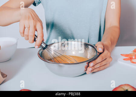 Frau Hände Schneebesen Eier in Metall Schüssel für Apfelkuchen. Close-up Frauen sitzen auf dem Tisch kochen Obst Kuchen. Stockfoto