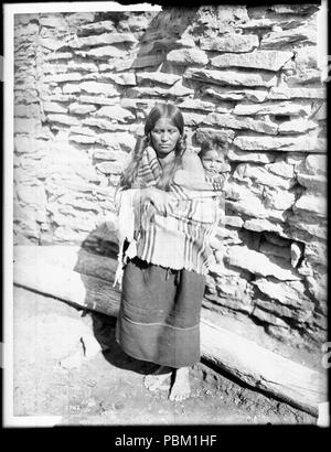 . Englisch: Hopi Indianer Mutter ihr Baby auf dem Rücken außerhalb einer nativen Steingebäude, Ca.1900 Foto: Hopi Indianer Mutter ihr Baby auf dem Rücken außerhalb einer nativen Steingebäude, Ca. 1900. Ihr dunkles Haar ist in engen Zöpfen an den Seiten des Kopfes getragen. Sie trägt eine gestreifte Decke über die Schultern über ihren Knöchel - Länge Kleid. Das Baby ist umschlungen in die Decke auf ihrem Rücken. Direkt hinter der Frau barfuss legt eine lange Holz. Rufnummer: CHS-3742 Fotograf: Pierce, C.C. (Charles C.), 1861-1946 Dateiname: CHS-3742 Abdeckung Datum: ca. 1900 Teil der Sammlung: Kalifornien Hi Stockfoto