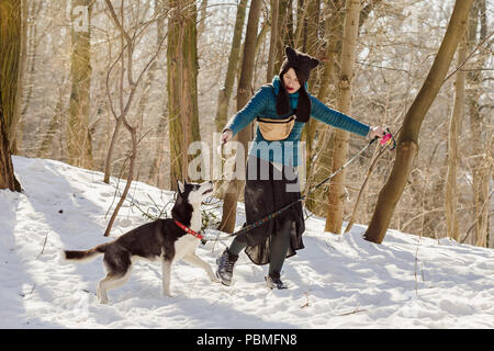 Stilvolle Frau mit lustigen Hut und Rock mit ihr spielen Siberian husky Hund im Winter Wald mit dem Stick Stockfoto