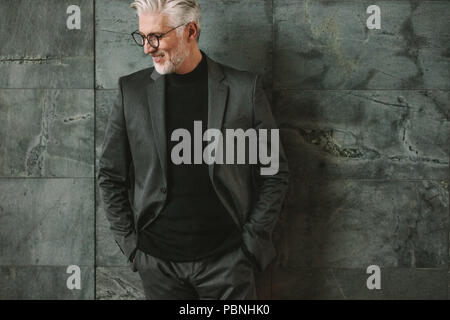 Portrait von Stattlichen reife Geschäftsmann stehend gegen graue Wand. Stilvolle älterer Mann, der mit den Händen in der Tasche und Wegsehen. Stockfoto