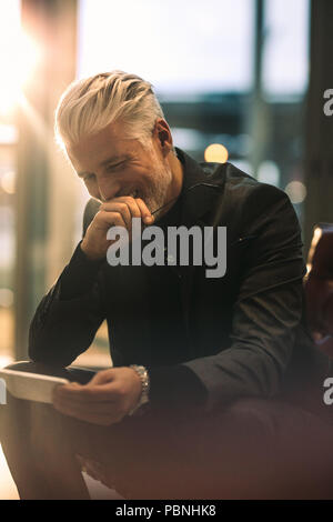 Vertikale Portrait von Reifen Geschäftsmann bei seinem Telefon suchen und lächelnd. Glücklich Mann in formelle Kleidung im Büro Lobby mit Smartphone sitzen. Stockfoto