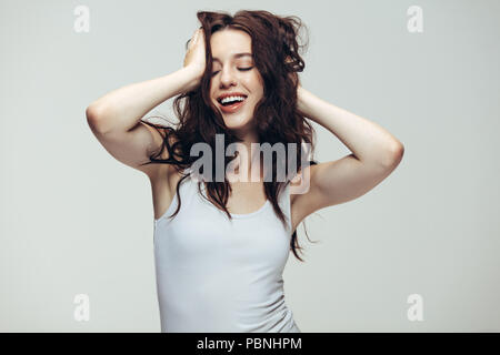 Porträt der glückliche junge Dame in Weiß stehend, mit ihren Händen in Haar vor grauem Hintergrund. Lächelnde Frau in Weißem tank top posiert im Studio. Stockfoto