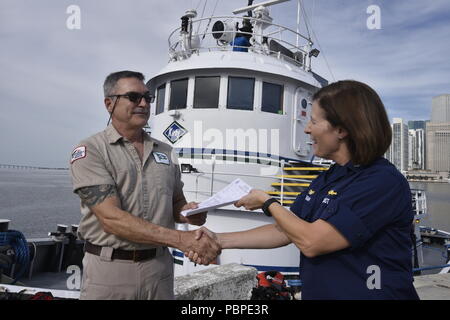 Kapitän Megan Dean, der Kommandant der Küstenwache Sektor Miami, ein Zertifikat der Inspektion Kapitän Steven Johnson, Kapitän der tugboat Miami im Hafen von Miami, 20. Juli 2018. Die Zertifikate der Inspektion auf den Schlepper Peggy Ann, Jean Ruth und Miami ausgestellt sind das Ergebnis von Jahren der Arbeit zwischen Ihnen und der Küstenwache zu schaffen Sicherheit Vorschriften für die Inspektion Normen und Safety Management Systems von Abschleppen von Schiffen. Coast Guard Foto von Petty Officer 3. Klasse Brandon Murray. Stockfoto