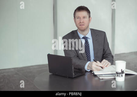 Kaukasier Männlich in seinen 30 trägt einen grauen Anzug, der Arbeit in einem aufgeräumten Schreibtisch, mit einem kleinen Laptop, Notebooks und einen großen Kaffeebecher. Stockfoto