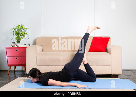 Schönen kaukasischen brünette Frau in schwarzer Kleidung auf Blau yogamat Yoga Körperhaltung nach unten liegend mit erhöhten am linken Bein. Stockfoto