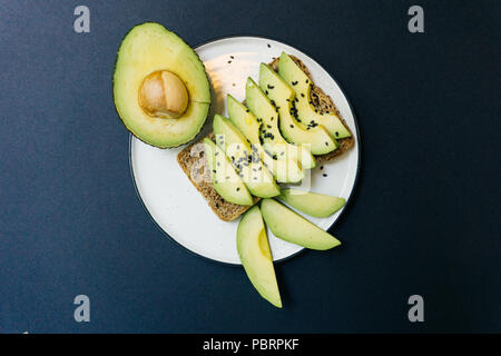 Toast mit Avocado in Scheiben geschnitten und schwarzem Sesam auf Vollkornbrot. Weiße Platte auf schwarzem Hintergrund Stockfoto