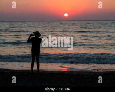 Ein Mann, der ein wenig Hut Fotografien den Sonnenuntergang am Strand im Urlaub Stockfoto