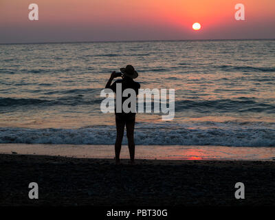 Ein Mann, der ein wenig Hut Fotografien den Sonnenuntergang am Strand im Urlaub Stockfoto