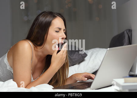 Müde Frau gähnen mit einem Laptop auf einem Bett in der Nacht zu Hause Stockfoto