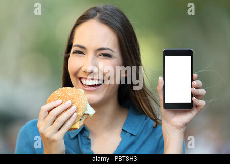 Gerne Frau mit einem Hamburger zeigt ein Bildschirm "Telefon" auf der Straße Stockfoto