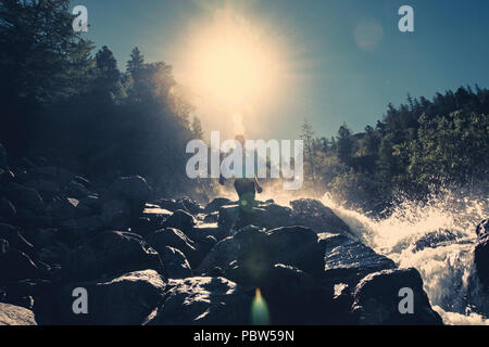 Ein Mann steht auf einem Stein vor einem Wasserfall unter der hellen Sonne. Wasserfall im Wald. ein Spray von Wasser. Stockfoto