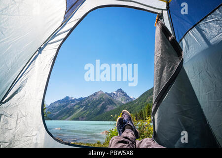 Füße Kletterer Mann entspannende Beobachten von einem Zelt Camping outdoor, Reisen Lifestyle Konzept am Ufer eines Bergsees Schnee Berge im Hintergrund Stockfoto