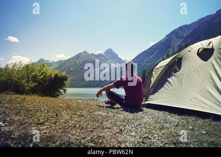 Allein man Reisen in der Nähe von dem See Berg Hintergrund, Konzept alleine reisen. Junge tourist Guy sitzt nach einem grünen Zelt und schauen an einem schönen Moun Stockfoto