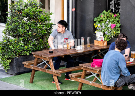 London, Großbritannien, 22. Juni 2018: Menschen auf dem Bürgersteig Straße in England sitzt, durch Sawyers Arms Pub trinken Pint Bier im Glas mit einem jungen asiatischen Menschen, Stockfoto