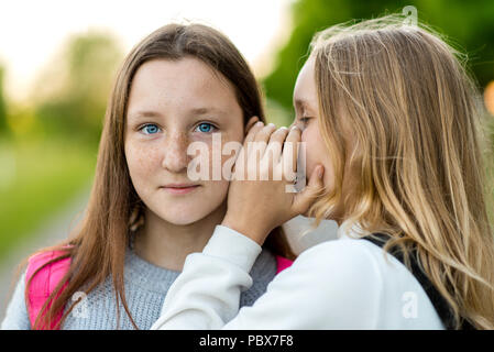 Zwei Mädchen Schülerin, im Sommer in einem Park in der Natur. Sie flüstert in mein Ohr. Das Konzept, das Geheimnis, die Überraschung und die Wahrheit. Emotionen lächeln Genuss. Schönen blauen gzal und Sommersprossen im Gesicht. Stockfoto