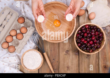 Zubereitung von hausgemachtem Süßgebäck mit Kirschen. Eier, Mehl und Beeren auf Holzgrund. Weibliche Hände teilen Eier. Speicherplatz kopieren. Stockfoto