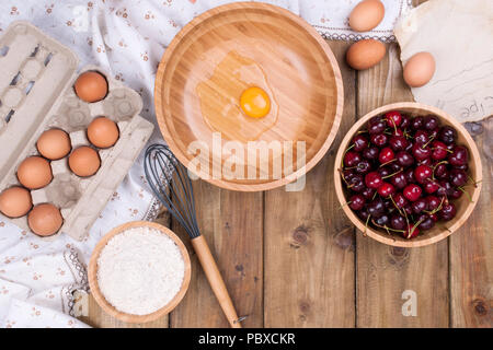 Zubereitung von hausgemachtem Süßgebäck mit Kirschen. Eier, Mehl und Beeren auf Holzgrund. Gehacktes Ei in einer Holzschüssel. Speicherplatz kopieren. Stockfoto