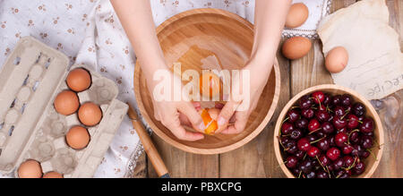 Zubereitung von hausgemachtem Süßgebäck mit Kirschen. Eier, Mehl und Beeren auf Holzgrund. Weibliche Hände teilen Eier. Speicherplatz kopieren. Stockfoto