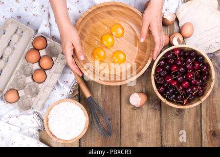 Zubereitung von hausgemachtem Süßgebäck mit Kirschen. Eier, Mehl und Beeren auf Holzgrund. Weibliche Hände teilen Eier in eine Holzschale. Speicherplatz kopieren. Stockfoto