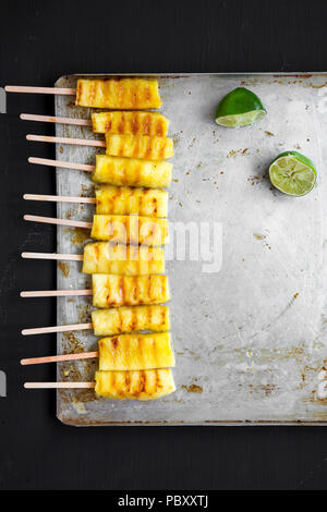 Gegrillte Ananas auf Bambusstäbe mit Kalk auf Fach über schwarzen Tabelle, Ansicht von oben. Von oben, flach und Overhead. Stockfoto