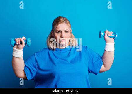 Porträt einer schweren übergewichtigen Frau mit Hanteln in Studio auf einem blauen Hintergrund. Stockfoto