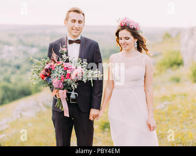 Der lächelnde jungvermählte Paar mit geschlossen Augen halten sich an den Händen. Der Bräutigam hält den Brautstrauß. Die horizontale Nahaufnahme. Stockfoto