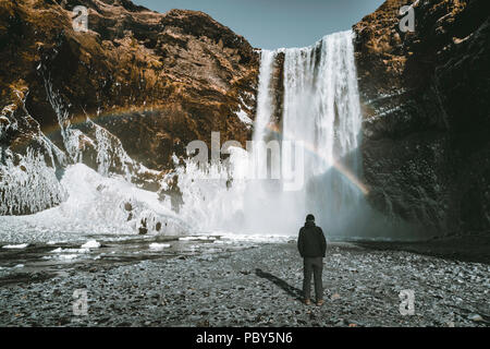 Eine Person admirnig die Schönheit der Skogafoss Wasserfall mit Regenbogen in Island entfernt. Stockfoto