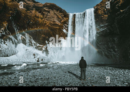 Eine Person admirnig die Schönheit der Skogafoss Wasserfall mit Regenbogen in Island entfernt. Stockfoto