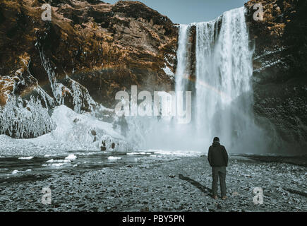 Eine Person admirnig die Schönheit der Skogafoss Wasserfall mit Regenbogen in Island entfernt. Stockfoto