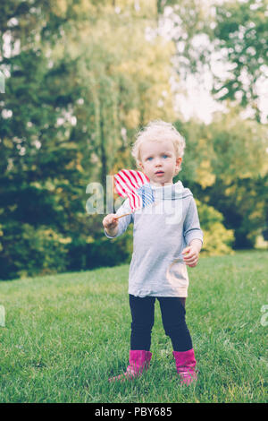 Happy adorable kleine blonde Kaukasischen girl Holding und wehende amerikanische Flagge in Park außerhalb Feiern 4. Juli Unabhängigkeitstag Flag Tag Konzept, ac Stockfoto