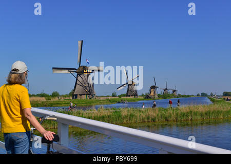 Radfahrerin, die auf der Unesco-Weltanlage Kinderdijk, Südholland, Niederlande, die berühmten Windmühlen neben Kanälen betrachtet Stockfoto