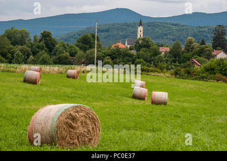 Kroatien, Plitvicer: gelb Heuballen mit grünen Wiesen auf Ackerland in der Landschaft und die Pfarrkirche von der kleinen Ortschaft Drežnik Grad Stockfoto