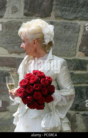 Die blonde Braut mit dem bridal Tasche und roten Rosen in der Hand entspannt mit einem Glas Champagner nach dem Empfang der Gäste. Stockfoto