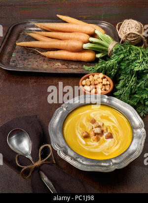 Leckere vegetarische Karotten Suppe serviert mit Sahne Croutons in Metall Zinn Schüssel. Bündel von Karotten, Croutons und Löffel auf dunklen braunen Hintergrund Stockfoto