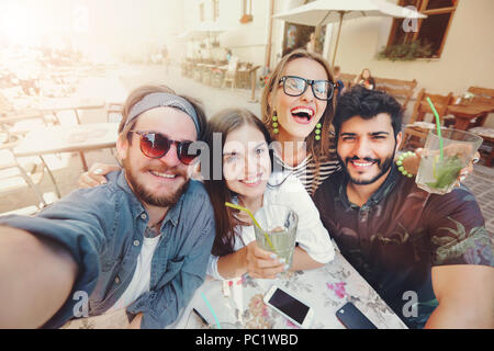 Vier fröhliche fröhliche Freunde eine selfie beim zusammen im Cafe sitzen und zeigen mohito im Freien Stockfoto