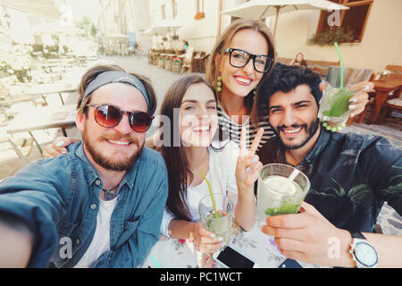 Vier fröhliche fröhliche Freunde eine selfie beim zusammen im Cafe sitzen und Frieden Geste im Freien Stockfoto