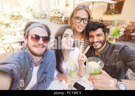 Vier fröhliche fröhliche Freunde eine selfie beim zusammen im Cafe sitzen und Frieden Geste im Freien Stockfoto