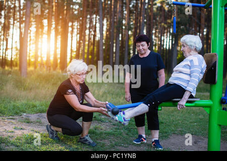 Eine ältere Frau auf einer Sport Simulator im Park, zwei erwachsene Frauen helfen, ihre Übungen zu tun. Health Club und Rehabilitation. Stockfoto