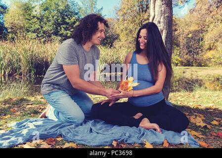 Junger Mann, der Blätter im Herbst zu seiner schwangeren Frau auf dem Boden sitzend. Selektiver Fokus Stockfoto