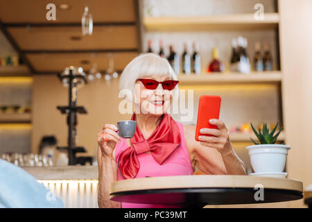 Lieblingsgetränk. Positive schöne Frau trinkt Espresso, während Sie an Ihrem Bildschirm des Smartphones Stockfoto