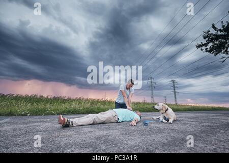 Reanimation auf der Landstraße gegen Sturm. Treuer Hund (Labrador Retriever) in der Nähe von seinem Besitzer warten. Themen Rettung, Hilfe und Hoffnung. Stockfoto